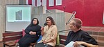 Drei Personen sitzen auf einer Bank, die mittlere spricht in ein Mikrofon. Im Hintergrund ist ein Foto auf einer Leinwand zu sehen und dahinter an der Wand weiß Stoffbahnen mit Namen.