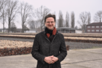 Oliver trägt einen Mantel und einen Schal, er hat eine Brille auf und blickt in die Kamera. Im Hintergrund die KZ-Gedenkstätte Neuengamme