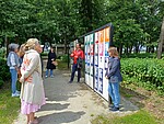 Menschen stehen vor einer Wand mit Postern, eine Person erklärt etwas, im Hintergrund Bäume.
