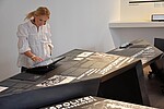 Eine Frau blickt interessiert auf einen Bildschirm, der auf der Ausstellungsplatte befestigt ist, bei der es um Zwangsarbeit geht.