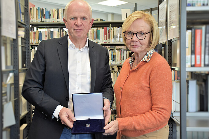 Zwei Personen halten ein Kästchen mit einem Ring und schauen in die Kamera. Das Foto wurde in der Bibliothek der KZ-Gedenkstätte Neuengamme aufgenommen.
