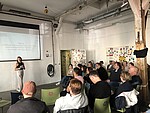 In einem Raum sitzen 25 Personen auf grünen Stühöen in einem Raum, an den Türen im Hintergrund kleben Aufkleber. Links steht eine Frau mit Moderationskarten in der Hand und spricht zum Publikum.