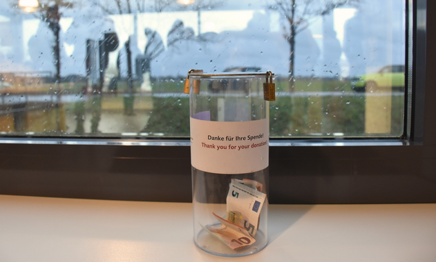 [Translate to Français:] Eine Spendenbox aus Plastik, darin Geldscheine. Aufkleber "Danke für Ihre Spende". Im Hintergrund ein Fenster mit Regentropfen.