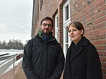Ansgar und Natascha stehen draußen und blicken in die Kamera, hinter ihnen eine Treppe und eine Backsteingebäudewand. Es liegt Schnee.