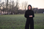 Eyleen trägt schwarze Kleindung und steht vor einer Wiese und Bäumen. Ihre Hände berühren einander.