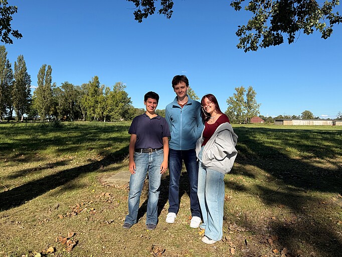 Josefa, Jack und Arina stehen auf dem Gelände der Gedenkstätte vor einem Feld