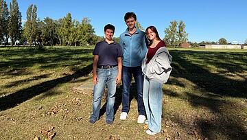 Josefa, Jack und Arina stehen auf dem Gelände der Gedenkstätte vor einem Feld