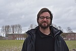 Ansgar Karnatz trägt eine Brille und eine schwarze Jacke. Ansgar lächelt in die Kamera, im Hintergund Wiese und Backsteingebäude.
