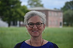 Dagmar Bonin trägt eine Brille und Ohrringe, sie blickt in die Kamera. Im Hintergund Wiese und Gebäude.