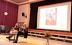 Eine Frau und ein Mann sitzen auf einer großen Bühne, im Hintergrund eine Projektion mit einem Schwarz-weiß-Foto.