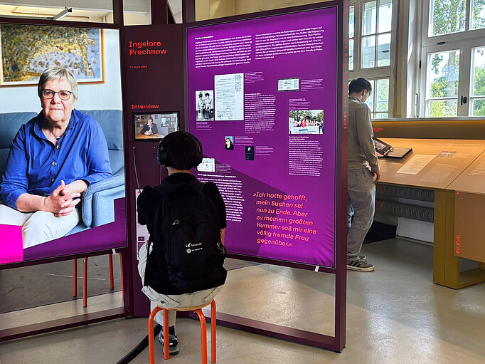 Ausstellung "Trotzdem da!" in der KZ-Gedenkstätte Neuengamme. Zwei Personen schauen sich die Ausstellung an. Ausstellungstafeln sind beleuchtet und zeigen ein großes Portrait sowie kleinere Dokumente