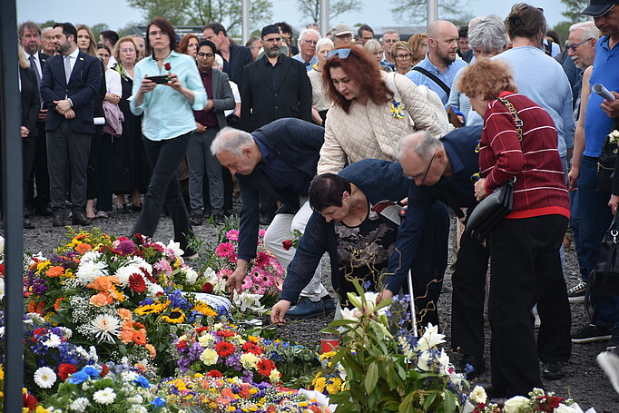Mehrere Personen legen Blumen ab, vor ihnen viele Blumen und Kränze, hinter ihnen weitere Personen, die zuschauen