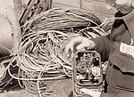 Eine schwarzweiß-Aufnahme, auf der viel Kabelgewirr zu sehen ist und im Vordergrund eine Hand, die ein einfaches selbstgebasteltes Radiogerät hält. Auf dem Ärmel des  Arms steht eine Nummer.