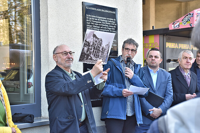 : Die Redner stehen vor dem Gebäude, einer hält ein historisches Foto in die Höhe, auf der das zerstörte Gebäude zu sehen ist, in dem 1944 das KZ-Außenlager eingerichtet wurde. 