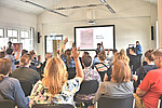 Personen sitzen auf Stühlen und schauen nach vorne, wo "How to Barcamp" auf einer Leinwand steht und 2 Personen mit Mikrofon in die Gruppe schauen
