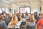 Personen sitzen auf Stühlen und schauen nach vorne, wo "How to Barcamp" auf einer Leinwand steht und 2 Personen mit Mikrofon in die Gruppe schauen