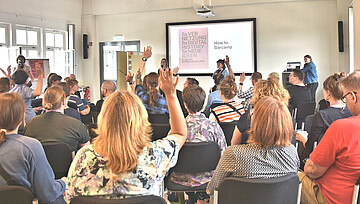 Personen sitzen auf Stühlen und schauen nach vorne, wo "How to Barcamp" auf einer Leinwand steht und 2 Personen mit Mikrofon in die Gruppe schauen