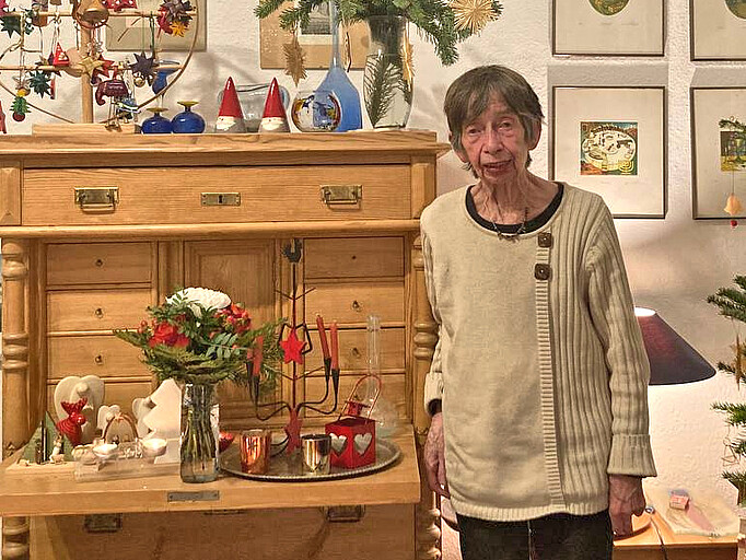 Eine ältere Dame steht vor einem Holzschrank mit Dekorationsobjekten