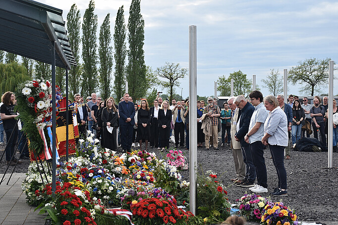 Kranzzeremonie am ehemaligen Arrestbunker mit vielen Blumengestecken, davor steht eine Gruppe von vier Personen, im Hintergrund weitere Personen.