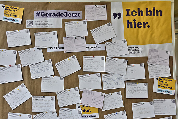 Postkarten sind an eine Pinnwand gepinnt mit Gründen, warum Menschen die KZ-Gedenkstätte Neuengamme besuchten. Dazu der Text "Ich bin hier"