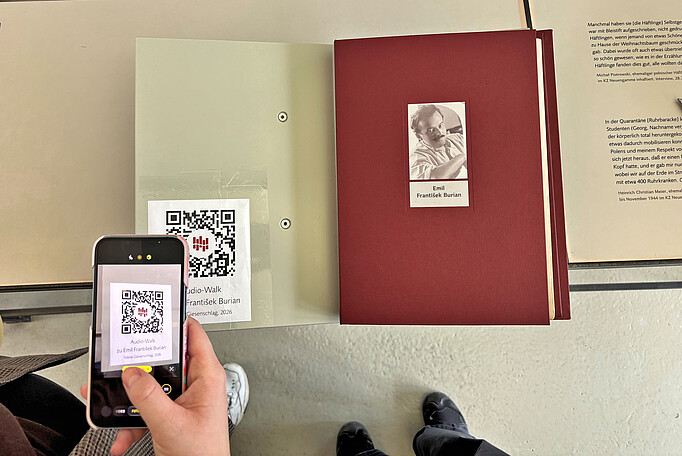 Rote Biografiemappe von Burian in der Ausstellung der KZ-Gedenkstätte Neuengamme, daneben ein Handy, welches einen QR-Code scannt