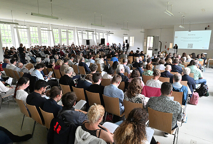 Gedenkveranstaltung in den ehemaligen Walther-Werken in der KZ-Gedenkstätte Neuengamme, viele Menschen auf Stühlen sind von hinten fotografiert, sie schauen auf eine