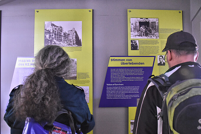 Eine Frau und ein Mann lesen sich die Ausstellungstafeln durch. Die Tafeln sind dunkelblau und hellgrün und hängen hochkant an der Wand. Auf der rechten Tafel ist die Überschrift „Stimmen von Überlebenden“ zu erkennen. 